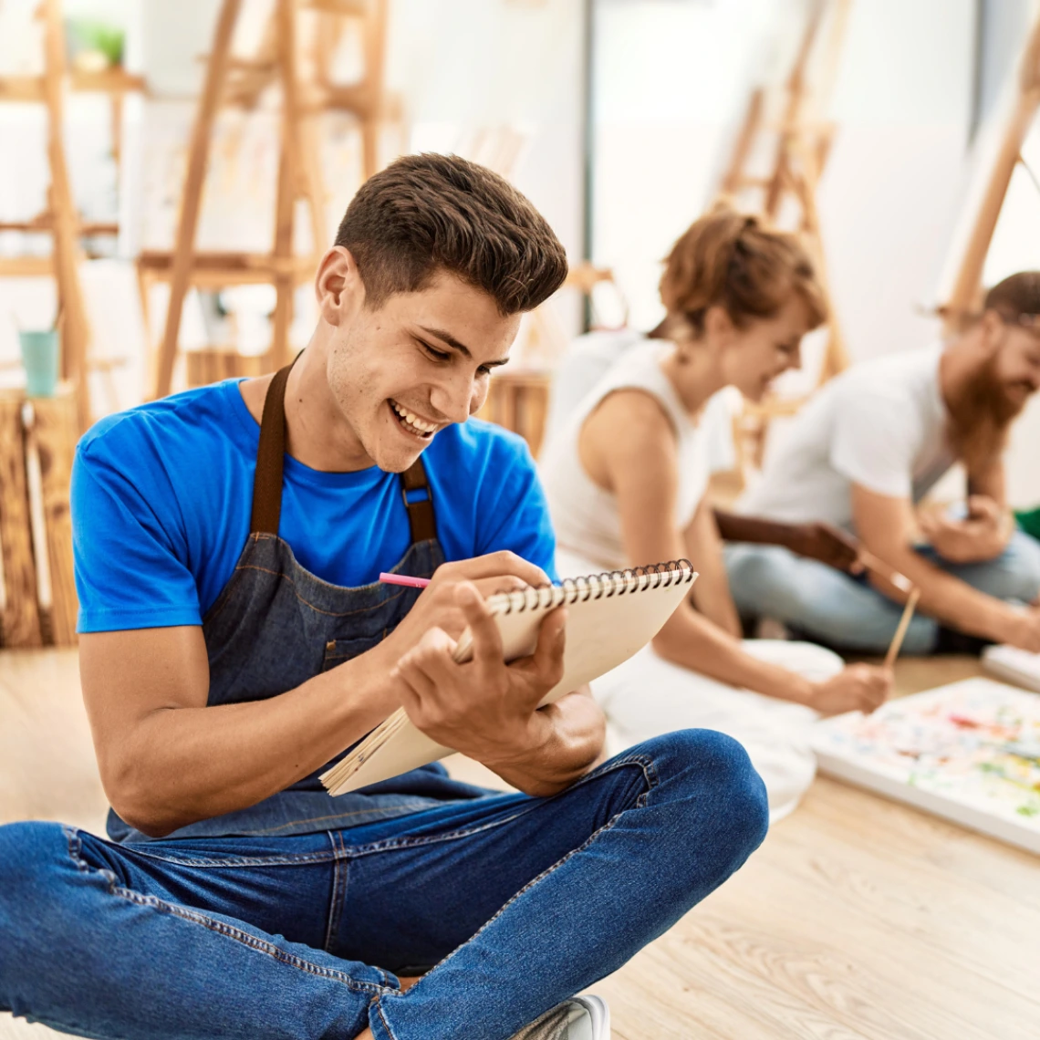 A group of people in an art studio drawing on the floor. In focus young man with a blue shirt and gray apron.